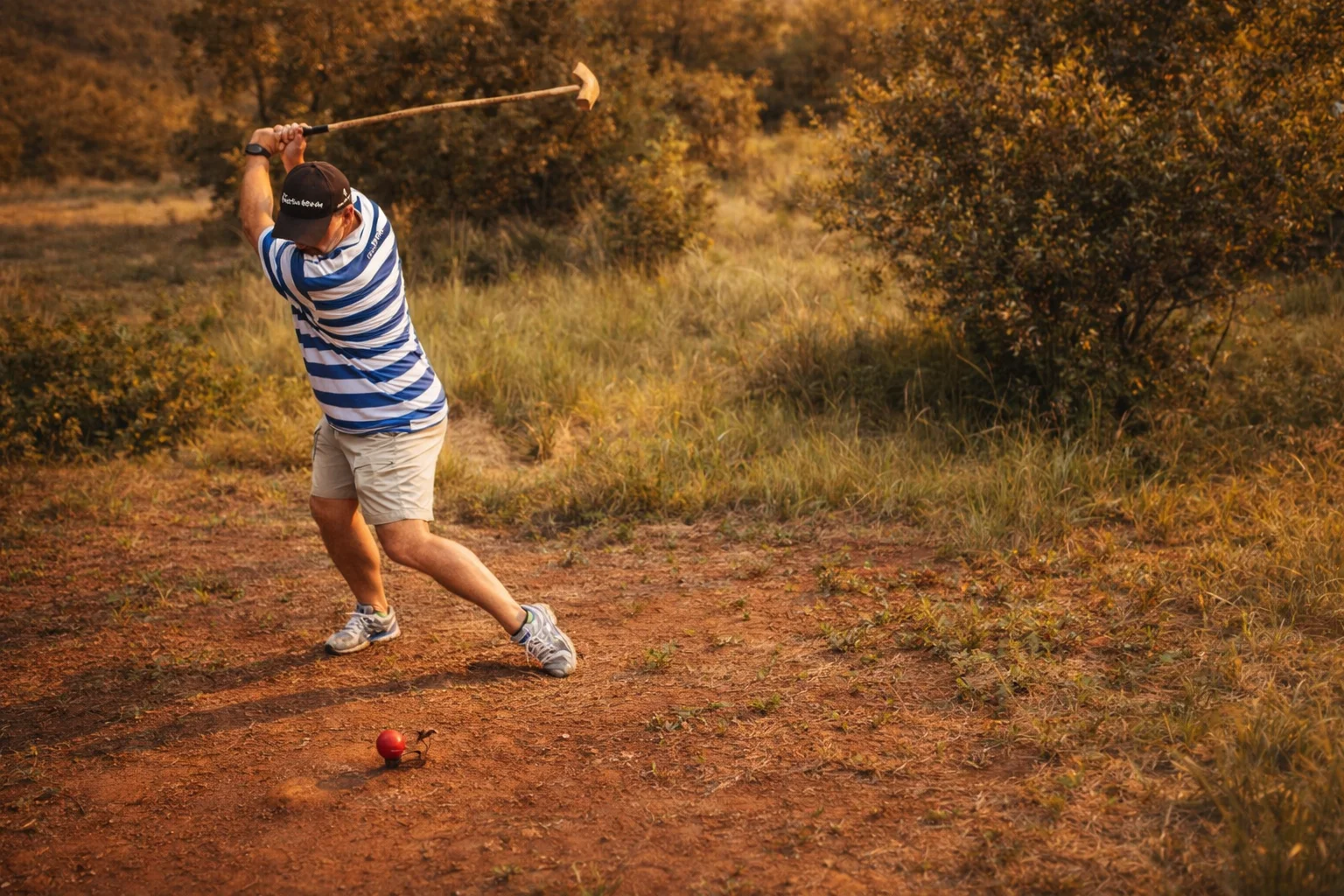 A player mid-swing with a handcrafted knobkierie on a bushveld Ancient Golf course at Shelter Rock in the Magaliesberg, golden afternoon light across the landscape.
