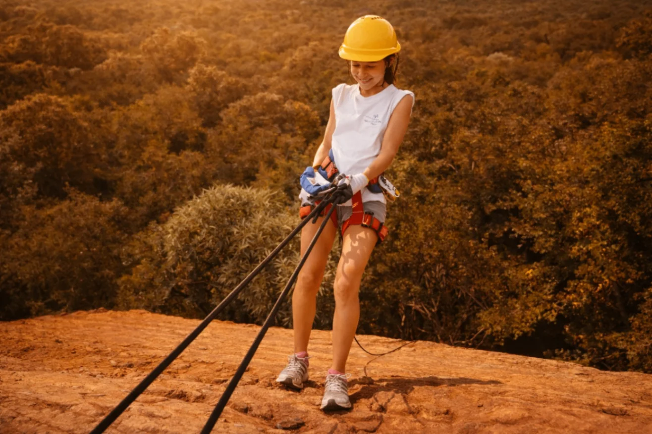 Abseiler on the edge of a Magaliesberg cliff preparing to lean back in a yellow helmet at sunset.